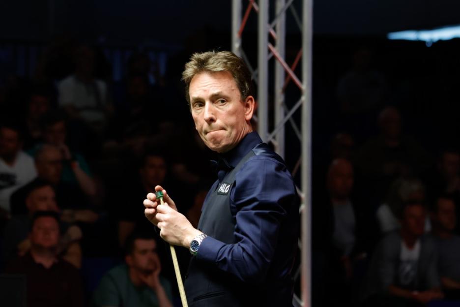 A male snooker player in a dark vest and shirt holds a cue stick, looking at the camera with a focused expression during the World Championship Qualifiers 2025.