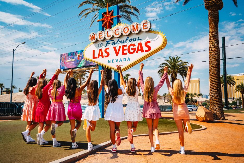 Nine women pose in front of the Welcome to Fabulous Las Vegas sign with their hands raised.