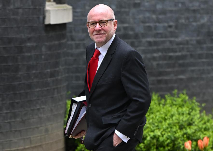 A bald man with glasses and a red tie carrying binders, smiles as he walks past a brick wall and green bushes.