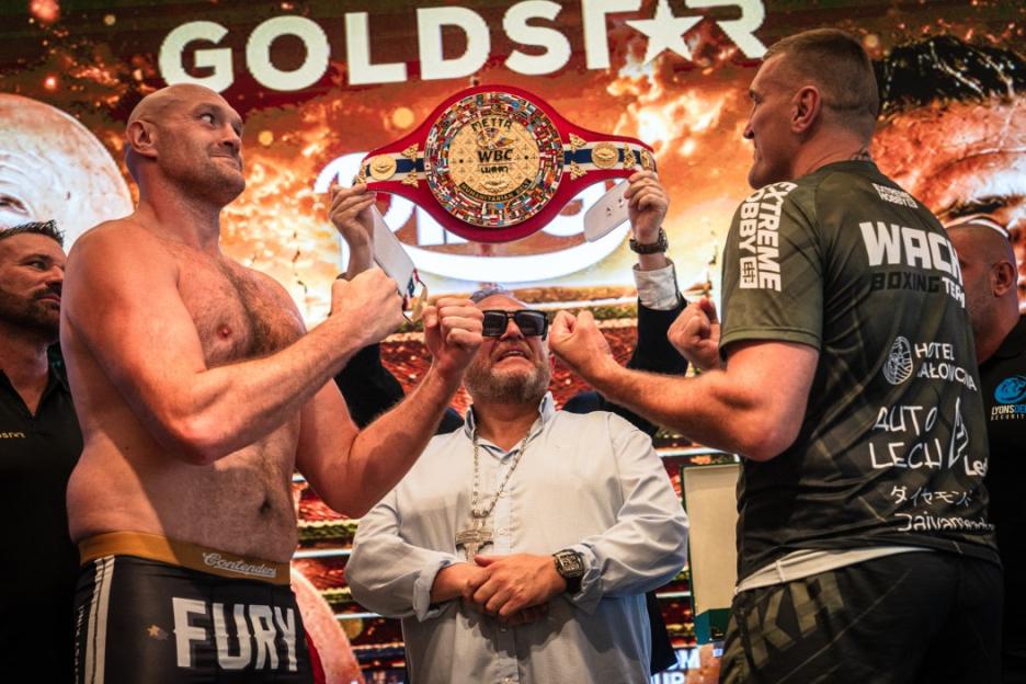 Boxers Tyson Fury and Mariusz Wach face off with a man in sunglasses holding up a championship belt between them.