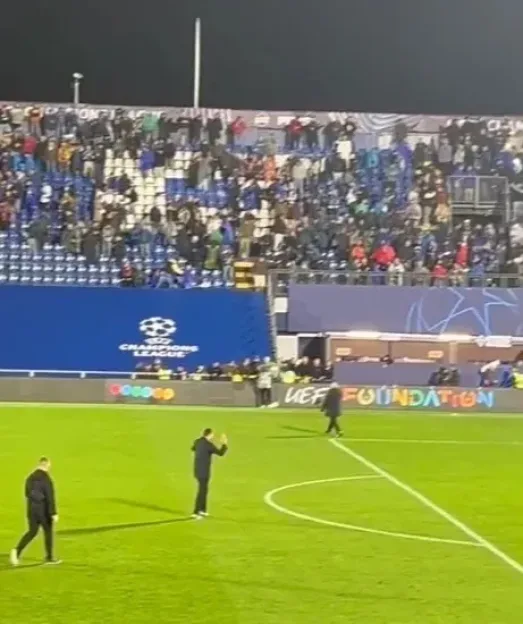 A soccer coach stands on the field, waving to a cheering crowd in the stadium.