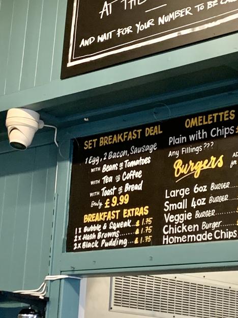 Menu board advertising breakfast deals, omelettes, and burgers, with a CCTV camera mounted on the wall above.