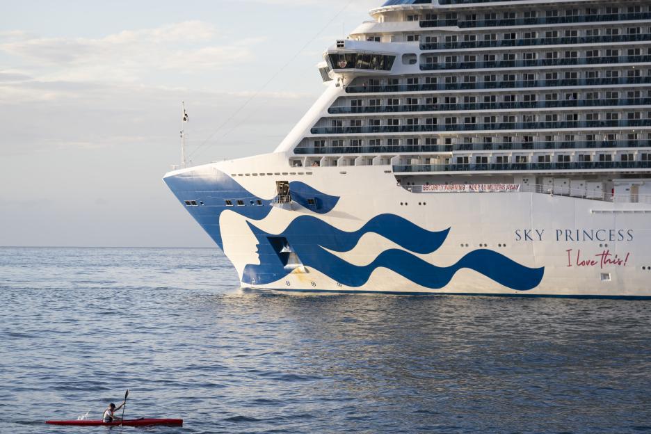 A person in a red canoe paddling in the sea next to the large Sky Princess cruise ship.