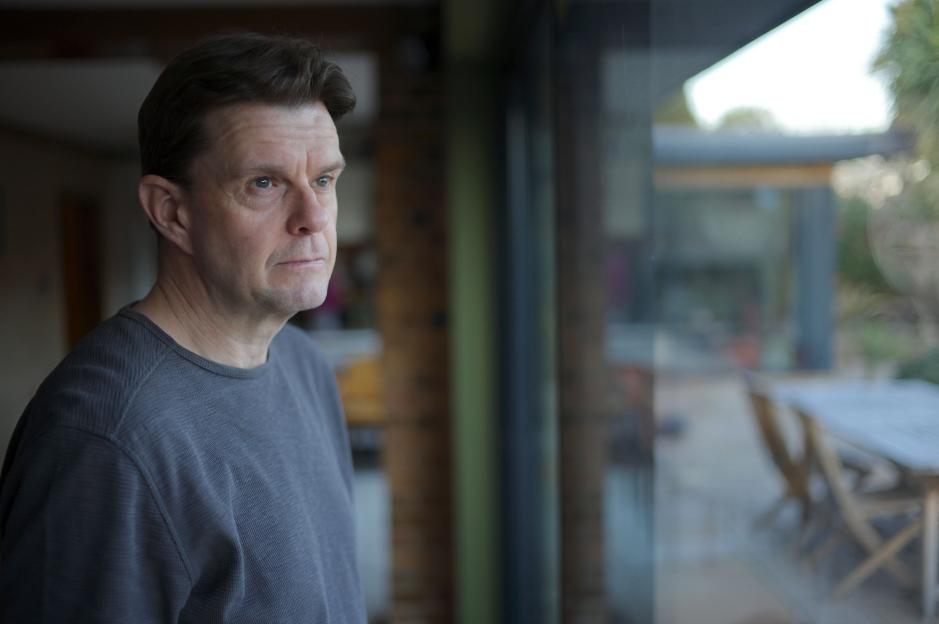 A man with a serious expression looks out a window, reflecting the sky and trees.
