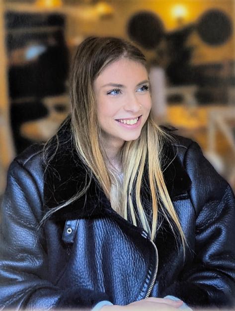 A young woman with long blonde hair, blue eyes, and a nose piercing smiles while wearing a black leather jacket.