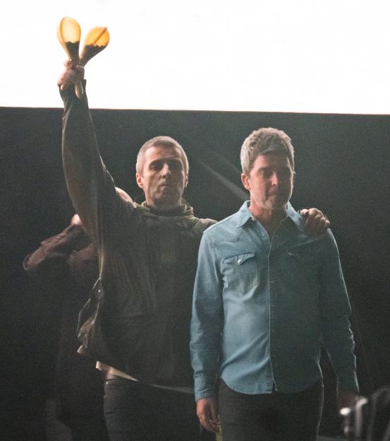 Liam Gallagher raising maracas with Noel Gallagher beside him on stage.