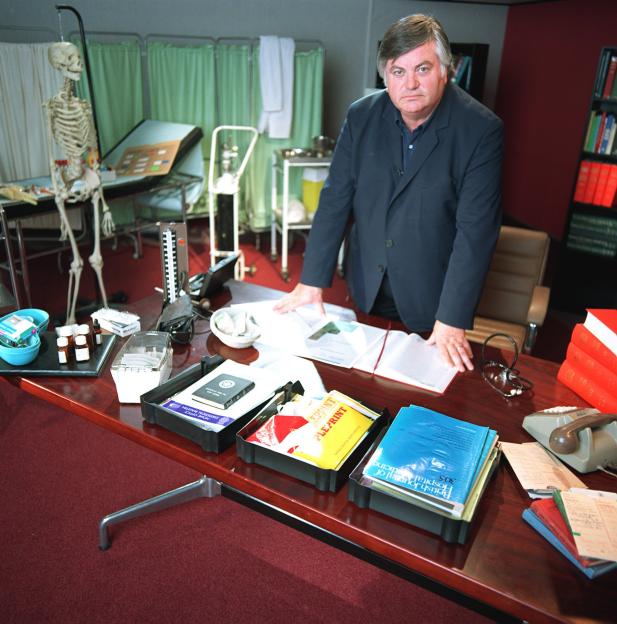 Roger Cook, a male TV presenter, stands behind a desk in a doctor's office, with a skeleton and medical equipment in the background.
