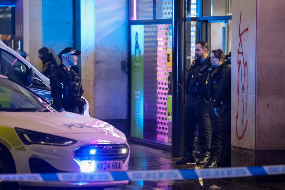Police officers and a police car behind blue and white police tape at night.