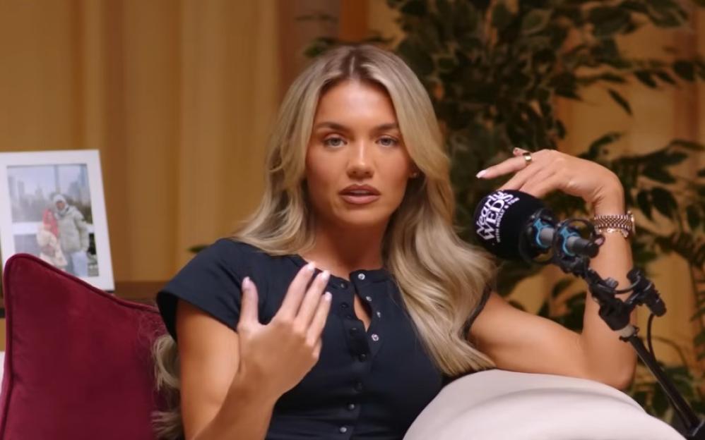 A woman with long blonde hair and a dark blue shirt speaks into a microphone during a podcast recording.