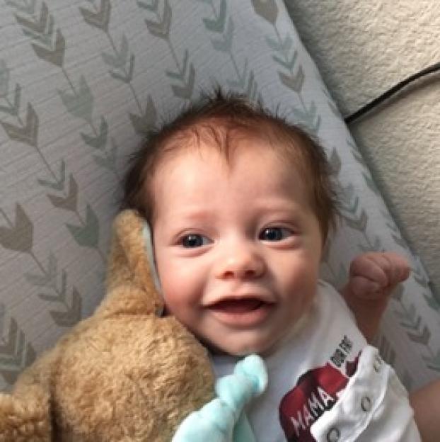 A close-up of a baby boy with brown hair and a slight smile, lying on a patterned pillow next to a stuffed animal.