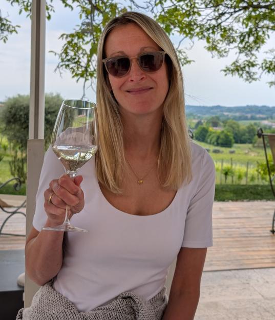 A woman in sunglasses holding a glass of white wine on a patio overlooking a vineyard.