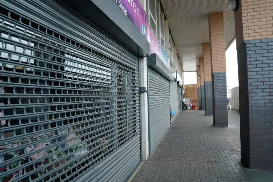 A row of closed shops with metal security shutters pulled down over their windows and doors, located along a paved walkway beneath a covered arcade.