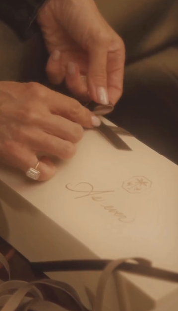 A woman's hands with a large emerald cut diamond ring on her left pinky finger tie a ribbon on a white gift box labeled "As Ever".