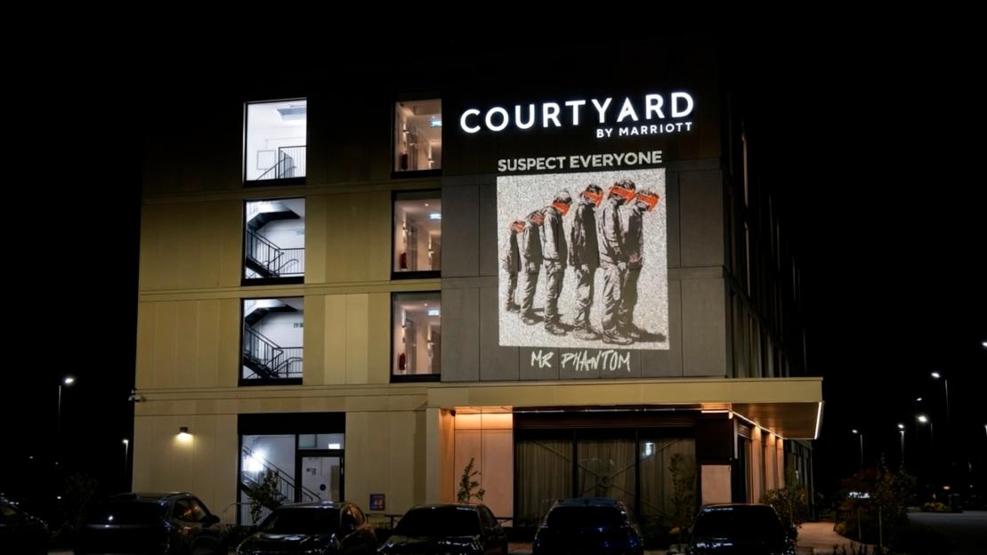 A projection of the words "SUSPECT EVERYONE" and an image of five figures with blindfolds and bound hands are displayed on the Courtyard by Marriott hotel building at night.