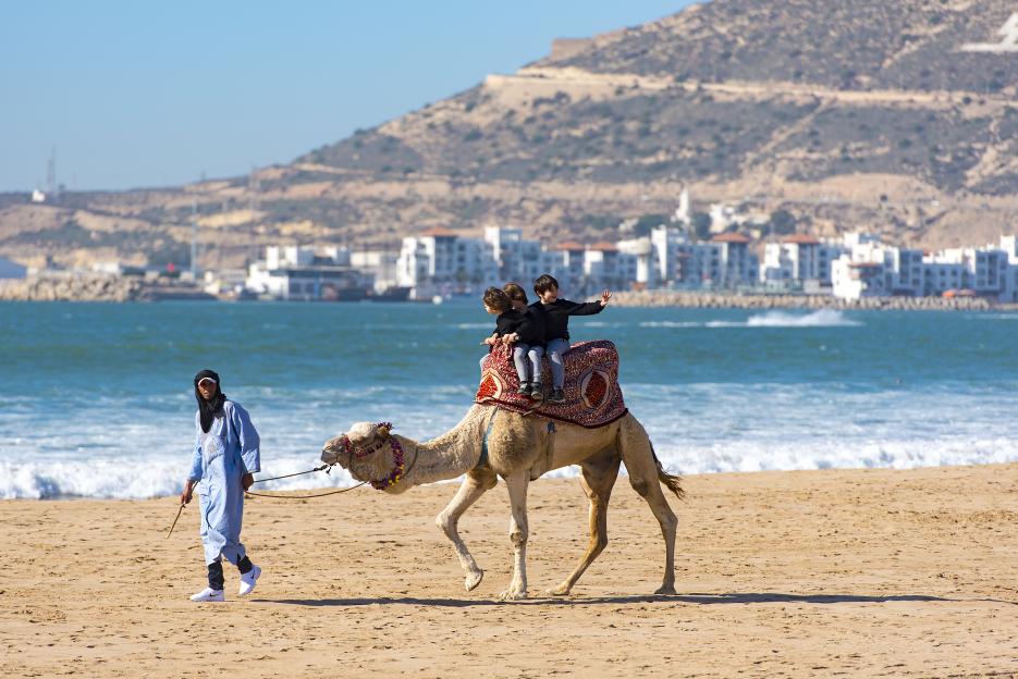 Two tourists riding a camel on a beach in Agadir, Morocco, led by a guide.