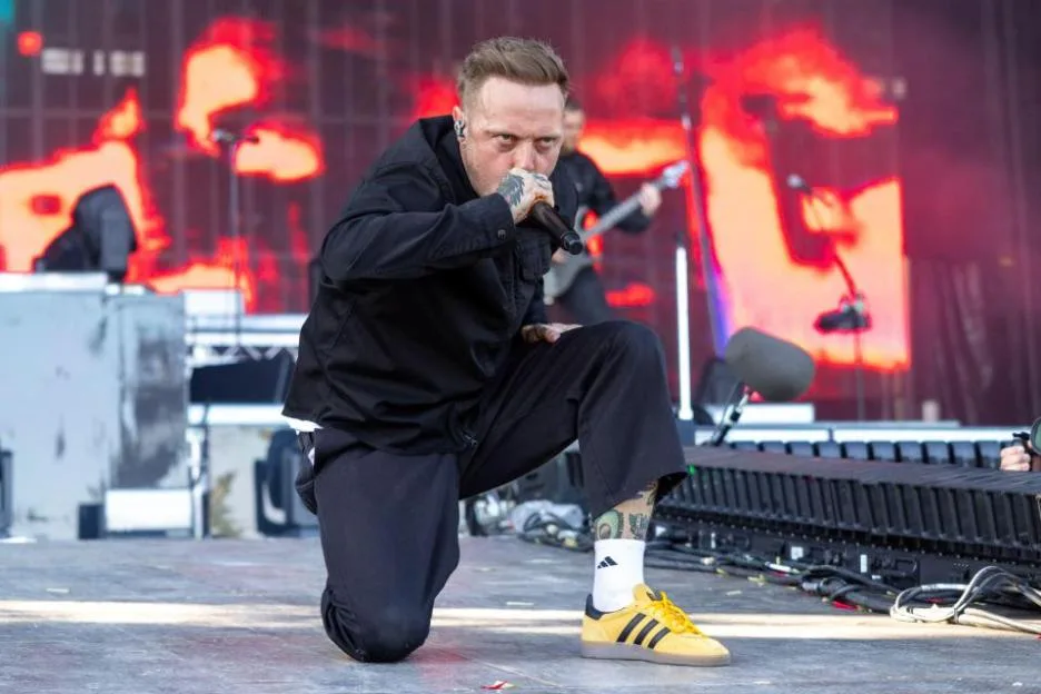 Sam Carter, singer of Architects, kneeling on stage and singing into a microphone with a red LED wall in the background.