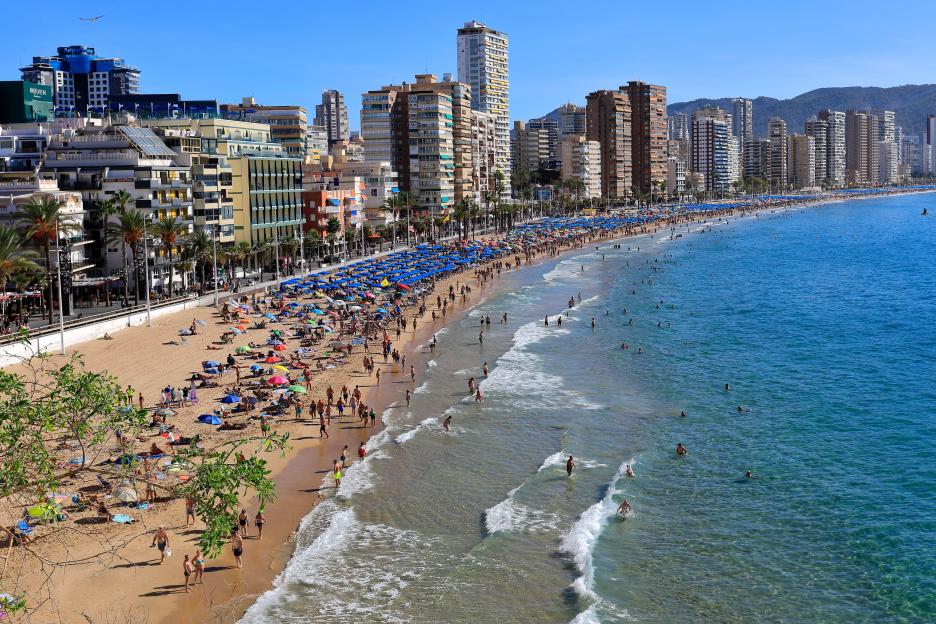 View over Levante beach in Benidorm town, Spain, showing tourists on the sand and in the water, with hotels and mountains in the background.