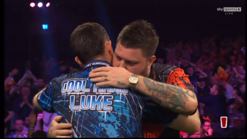 Luke Humphries, in a blue and black shirt with "COOL HANDS LUKE" on the back, embraces another man on stage after winning the 2025 PDC World Championship, while the crowd in the background cheers.