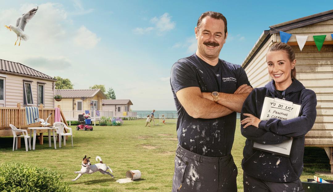 Two smiling people covered in paint splatters stand in front of a caravan park.