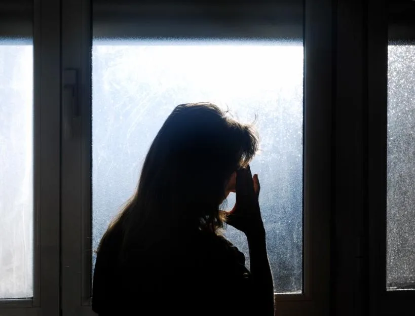 Woman in silhouette in front of a translucent window, hand on forehead in a sign of worry, anxiety, loneliness, abandonment