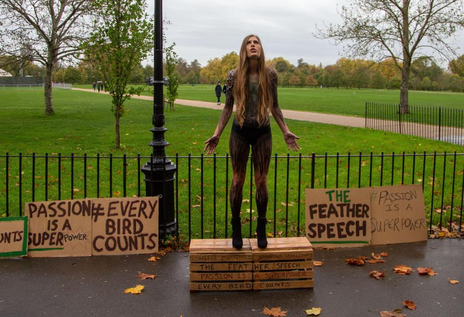 Conservationist Hannah Bourne-Taylor, body-painted to resemble a bird, stands on wooden crates in a park with protest signs for "The Feather Speech" campaign.