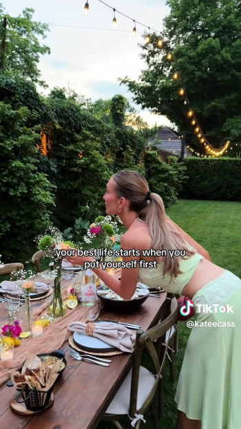 Woman in a green outfit placing flowers on an outdoor dining table, with text overlay "your best life starts when you put yourself first".