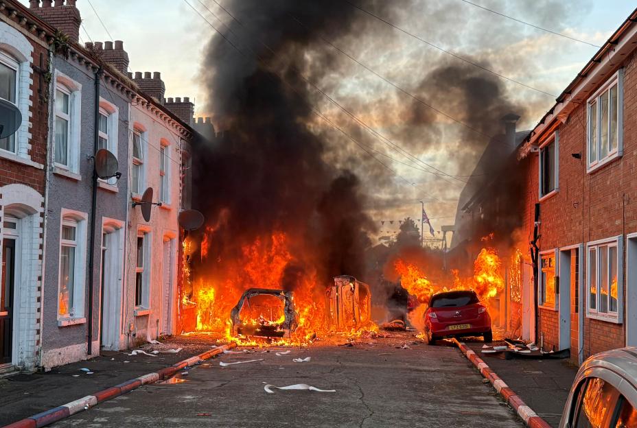 Cars ablaze on Lendrick Street in Belfast, with flames and smoke filling the street between two rows of houses.