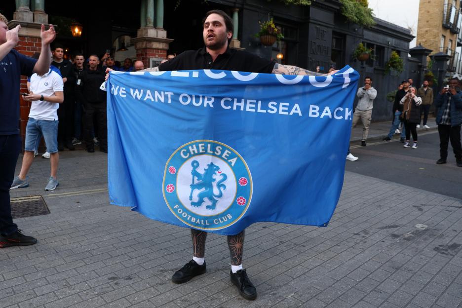 A Chelsea fan holds a banner that says "WE WANT OUR CHELSEA BACK" with the Chelsea Football Club logo.