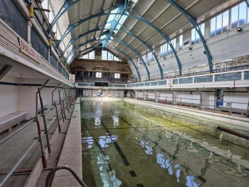 Indoor swimming pool with shallow, murky water and scaffolding on the sides, under a high arched ceiling with skylights.