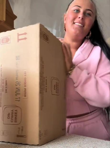 A woman in a pink sweatshirt holds a large cardboard box of curry sauce.