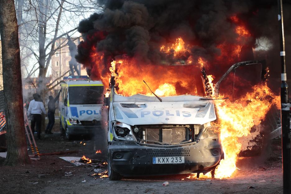 A police van on fire with large flames and dark smoke, with another police van visible behind it.