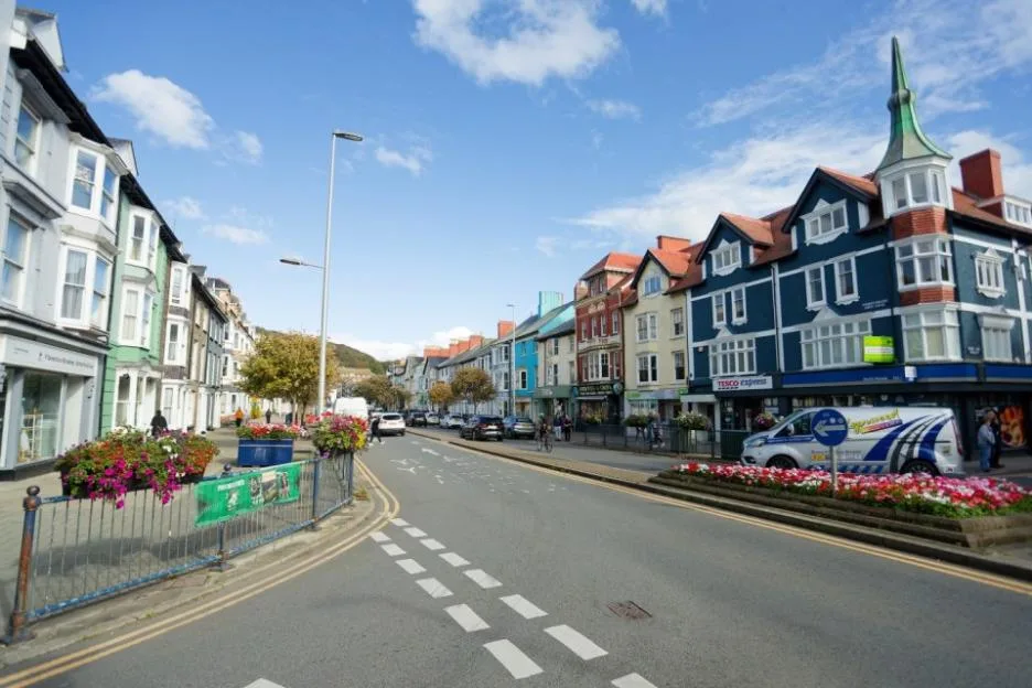 A street in Aberystwyth, Wales, UK, featuring historic buildings with colorful facades, shops, and a Tesco Express.