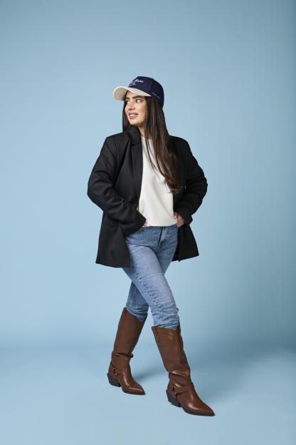 Emily Regan modelling a black blazer, white shirt, jeans, brown boots, and a blue cap.