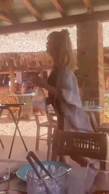 A blonde woman in a swimsuit and cover-up dances at a holiday resort, holding a large bottle of rosé.