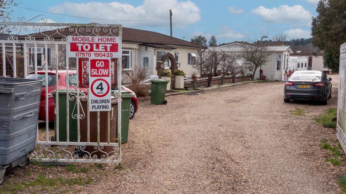 Gated entrance to the Buckles Lane traveler site with a "Mobile Home To Let" sign, speed limit warning, and houses and mobile homes visible inside.