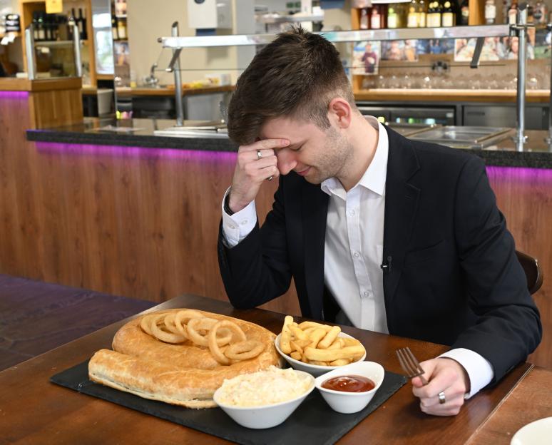Man at a restaurant attempts to eat a giant sausage roll with onion rings and fries.