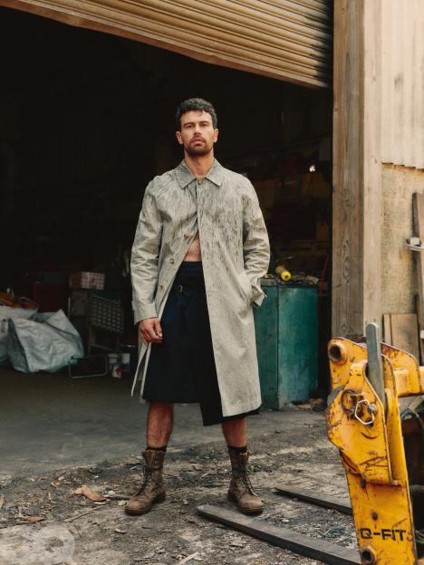 Theo James wearing a light-colored trench coat open over a dark kilt, with military boots, standing in front of an open garage.
