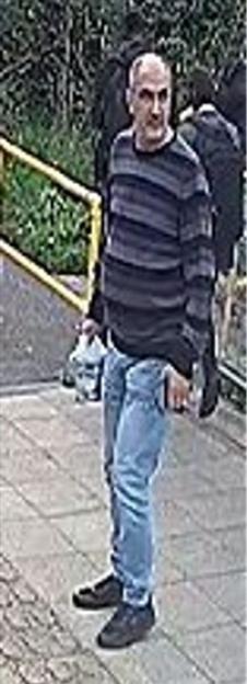 CCTV image of a man in a striped sweater, light blue jeans, and dark shoes, holding a plastic bottle and another item.