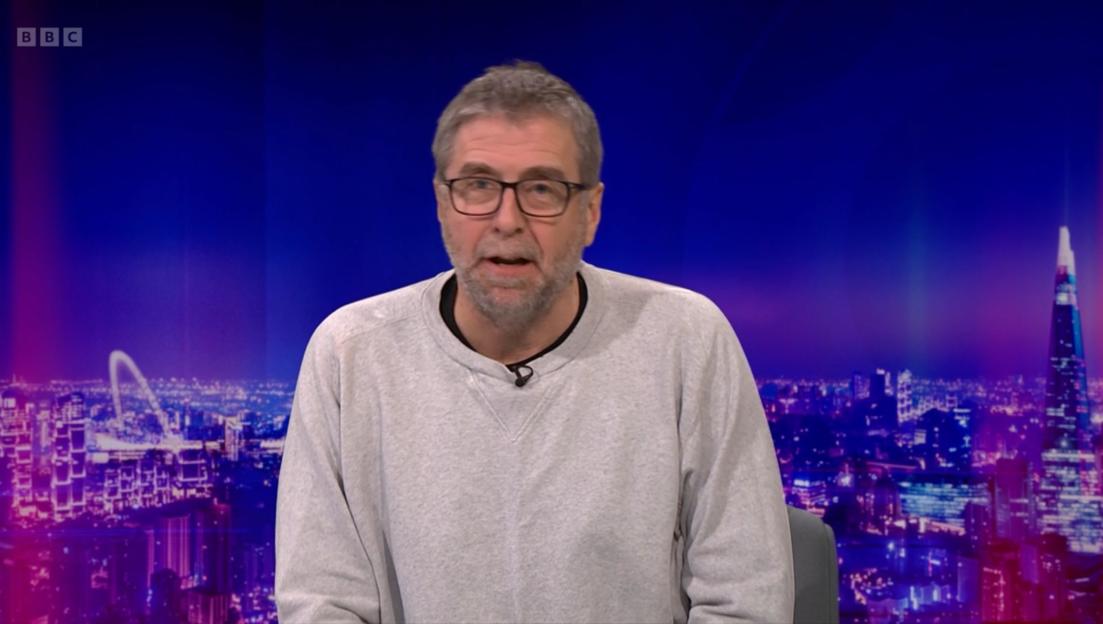 A man with glasses and a grey beard speaking on a BBC program with a city skyline at night in the background.