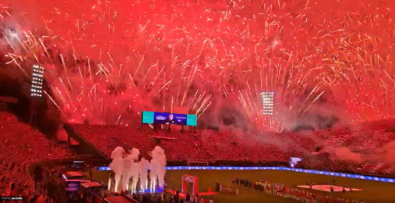 Red fireworks explode over a stadium filled with spectators.