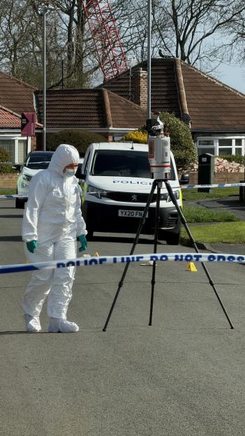 A forensic technician in a white hazmat suit and mask walks past a police van and a tripod-mounted camera at a crime scene.