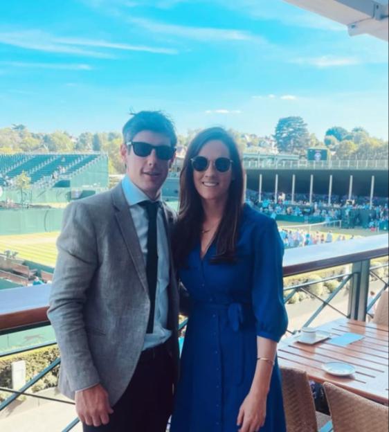 Two people standing on a balcony overlooking a tennis court.
