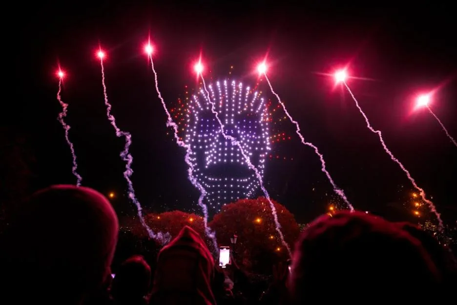 A crowd watching a drone display in the night sky, featuring a large illuminated face, framed by six bright pyrotechnic sparklers.