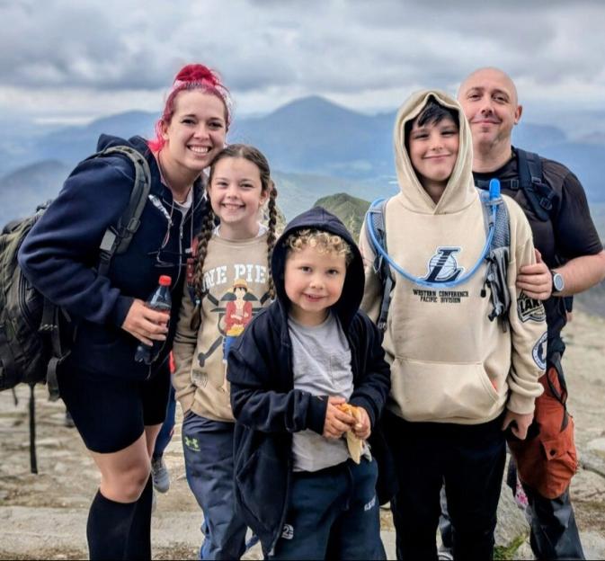 Chloe Wyman, her partner Aran Pierce, and his children Darcy, Bobby, and Edison pose together on a mountaintop.