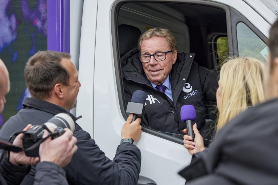 Harry Redknapp, wearing an Ocado jacket, sits in a delivery van while being interviewed by two people holding microphones.