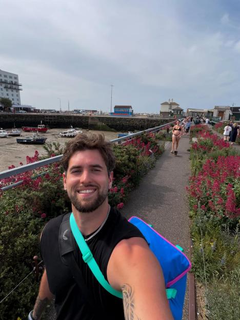Zach Noble in a selfie, carrying a bright blue and pink bag, with a harbor and promenade behind him.