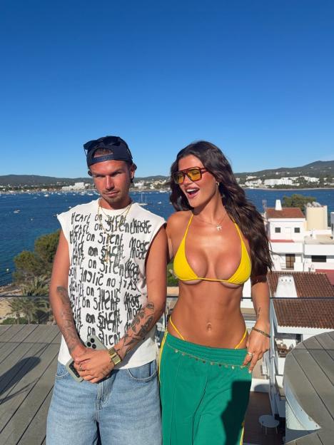 Matilda Draper in a yellow bikini with a man on a balcony overlooking the sea.