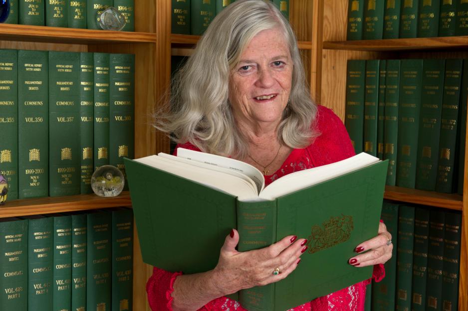 Ann Widdecombe, a woman with gray hair and a red top, smiles at the camera while holding an open "Parliamentary Debates Commons" book in front of a bookshelf filled with similar green books.