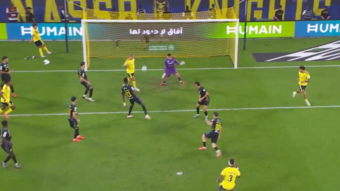 A football player in a black and yellow uniform kicks the ball towards the goal, defended by a goalkeeper and other players.