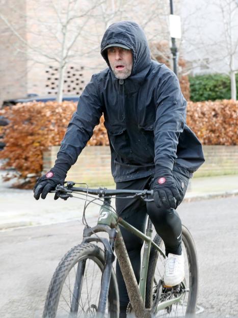Liam Gallagher riding a bicycle with a white beard and black hooded jacket.
